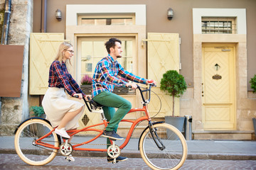Fototapeta premium Side view of active tourist couple in casual clothing, bearded man and blond woman in glasses cycling tandem bike along empty paved sidewalk by town residential houses.