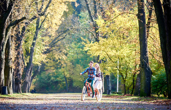 Young active tourist couple, handsome bearded man and attractive blond long-haired woman cycling together tandem double red bike by sunny alley with golden leaves on tall trees background in the fall