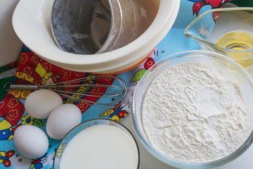 ingredients for pancakes are laid out on a multi-colored tablecloth, raw ingredients: eggs, milk, oil, flour