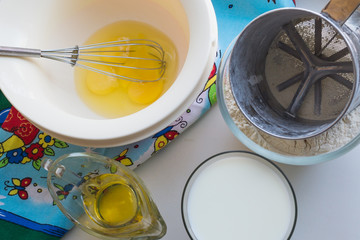 ingredients for pancakes are laid out on a multi-colored tablecloth, raw ingredients: broken eggs, milk, oil, flour