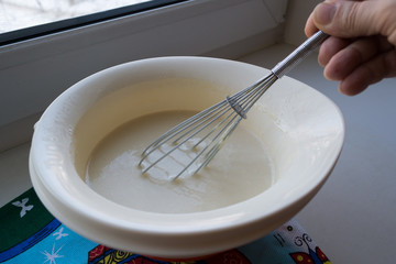 the finished pancake batter in a white bowl with a metal whisk holds the woman's hand of an elderly woman, dought