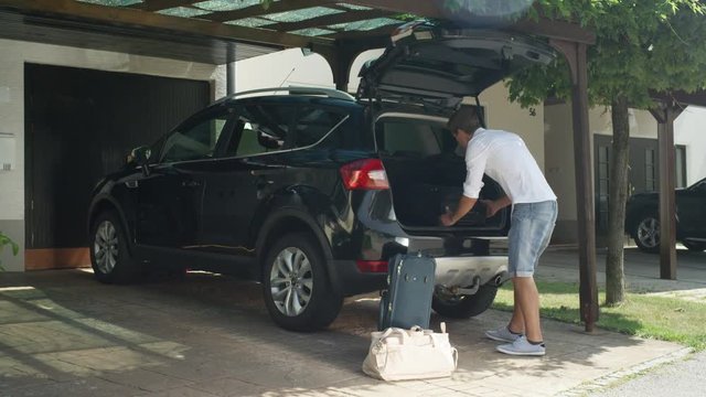LENS FLARE: Caucasian man yells in the sunny driveway at laughing girlfriend for packing too much luggage for their summer vacation. Young Caucasian newlywed couple overpacking before their honeymoon.