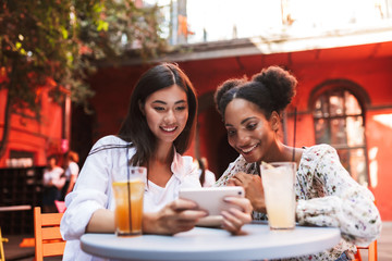 Young smiling ladies happily using cellphone together with drinks near on table while spending time in courtyard of cafe