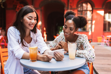 Two beautiful ladies happily using cellphone together with drinks near on table while spending time in courtyard of cafe