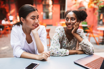 Young beautiful girls joyfully looking at each other while working with laptop together in cozy courtyard of cafe