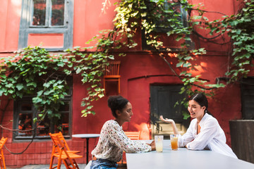 Pretty smiling girls dreamily talking while spending time together with cocktails in cozy courtyard of cafe