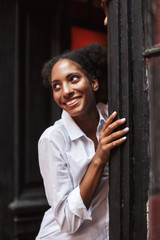 Fototapeta na wymiar Pretty joyful african girl with dark curly hair in white shirt happily looking aside while leaning on old black door