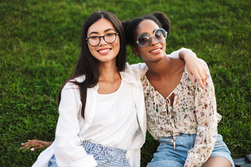 Two cheerful girls friendly hugging each other happily looking in camera while spending time together on grass in park