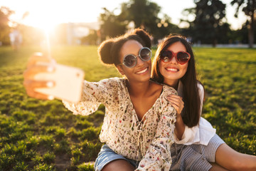 Pretty joyful girls in sunglasses sitting on grass happily taking photos on cellphone while...