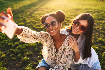 Beautiful smiling girls in sunglasses sitting on grass joyfully taking photos on cellphone while...