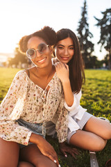 Naklejka premium Two pretty smiling girls sitting on grass joyfully looking in camera while spending time together in city park
