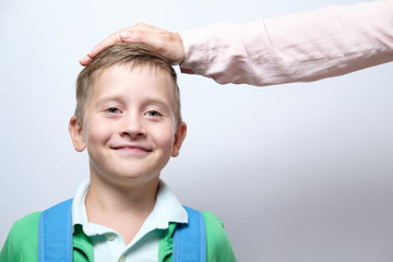Female hand on head of a cute schoolboy with blue backpack on a white background. concept of praise - obrazy, fototapety, plakaty