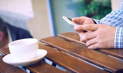 Man using a cell phone on cafe terrace and drinking coffee. Man drinks coffee. Man using cellphone. Mans hands with cell phone, coffee. Mans hands.