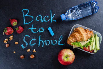 Back to school concept. Lunch box with croissant, fruits and vegetables on black background. Top view