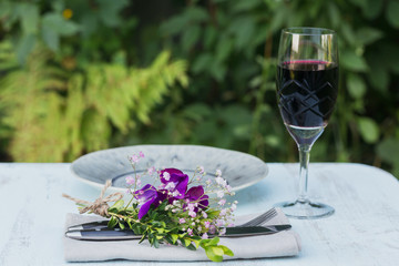 Rustic table setting with purple flowers and a glass of red wine on light wooden table.