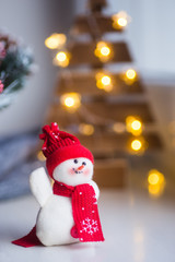 Closeup toy cute snowman in hat and scarf with warm garland lights on bokeh background.
