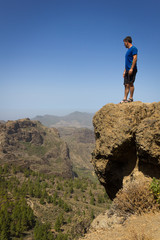 Young man on blue shirt looking down from top of cliff edge in Roque Nublo, Gran Canaria. Hiker on rocky mountain contemplating panoramic views. Outdoor activity, adventure, explore, freedom concepts