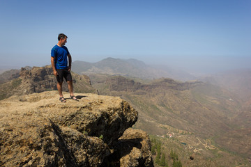 Fototapeta premium Young climber looking down from top of cliff edge in Roque Nublo, Gran Canaria. Adventurer on rocky mountain contemplating panoramic views. Outdoor activity, no fear, explore, freedom concepts