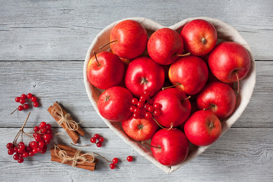Red apples in a heart-shaped basket, berries of viburnum and cinnamon on a wooden background