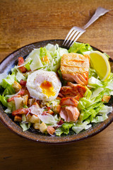 Salmon caesar salad. Crispy pan fried salmon fillet, bacon, poached egg, romaine lettuce and croutons in bowl on wooden table. vertical