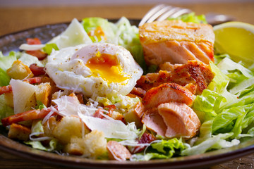 Salmon caesar salad. Crispy pan fried salmon fillet, bacon, poached egg, romaine lettuce and croutons in bowl on wooden table. overhead, horizontal