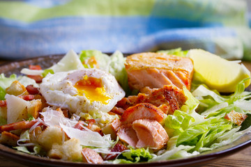 Salmon caesar salad. Crispy pan fried salmon fillet, bacon, poached egg, romaine lettuce and croutons in bowl on wooden table. horizontal