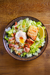 Salmon caesar salad. Crispy pan fried salmon fillet, bacon, poached egg, romaine lettuce and croutons in bowl on wooden table. overhead, vertical