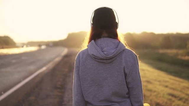 close-up portrait of a red-haired girl in headphones walking along the roadside at sunset. back view. slow motion.