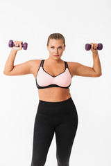 Fototapeta na wymiar Beautiful plump girl in sporty top and leggings holding dumbbells in hands while thoughtfully looking in camera over white background. Plus size model