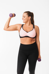 Fototapeta na wymiar Smiling plump girl in sporty top and leggings holding dumbbells in hands while happily looking aside over white background. Plus size model