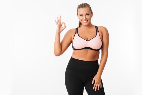 Smiling plump girl in sporty top and leggings happily showing ok gesture while looking in camera over white background. Plus size model
