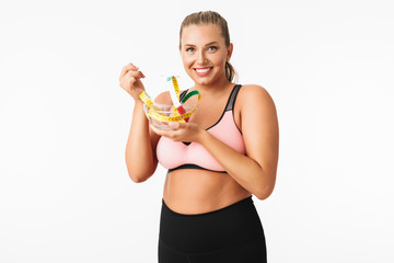 Young woman with excess weight in sporty top pretending that eat measuring tape from bowl while happily looking in camera over white background. Plus size model