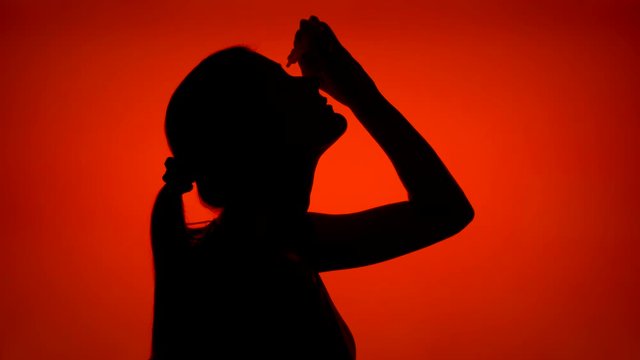 Silhouette of young woman applying eye drops. Female's face in profile putting water drops in her eyes on red background. Black contour shadow of teenager's half-face with medicine bottle