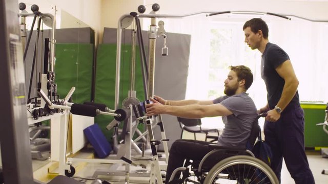 Young disabled man in wheelchair doing strength exercises for hands at the rehabilitation clinic. Doctor physiotherapist helping him