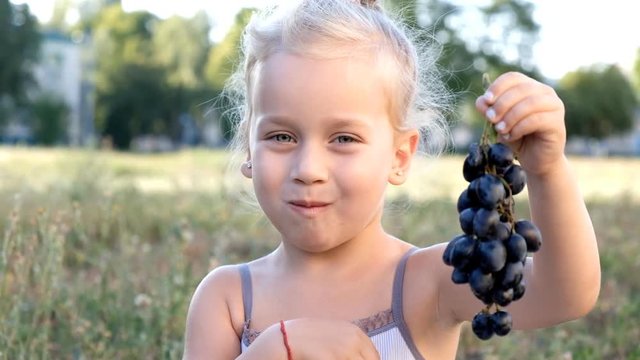 little blond girl sitting in the park on grass and eating grapes