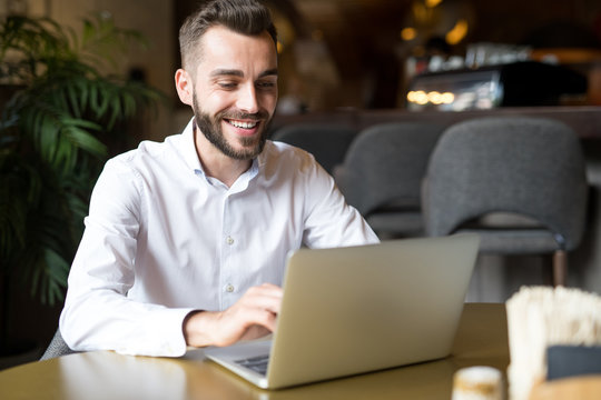 Portrait of handsome bearded businessman wearing white shirt smiling happily while using laptop sitting at table in cafe, copy space