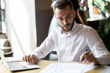 Portrait of handsome successful businessman wearing white shirt reading documents and using laptop while working at table in office or cafe, copy space