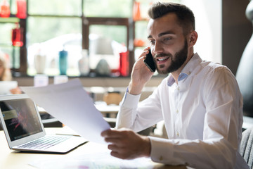 Portrait of handsome bearded businessman wearing white shirt speaking by phone and reading documents while working at table in office or cafe