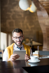 Portrait of smiling businessman wearing glasses using digital tablet while enjoying coffee break in bar, copy space