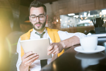 Warm toned portrait of handsome bearded businessman wearing glasses using digital tablet while enjoying coffee break in cafe behind glass wall in sunlight