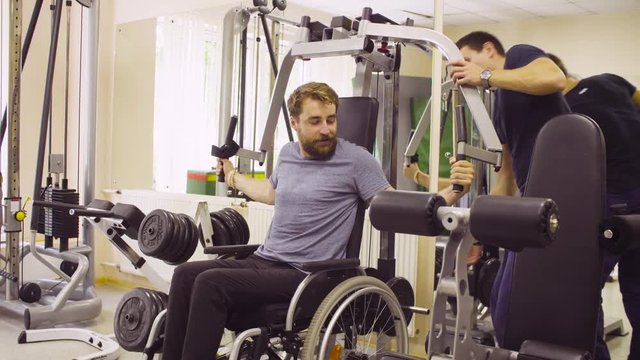 Young disabled man in wheelchair doing strength exercises for hands in the rehabilitation clinic. Doctor adding weight to the simulator. They talking and smiling.