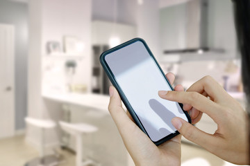 Female's hands holding mobile smartphone with white screen over blurred kitchen room background.
