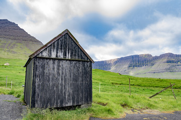 Fototapeta na wymiar Beautiful view of black faded wooden barn with green grass of sheeps farm and high mountain cliff in the background with cloudy weather sky in Faroe Islands rural