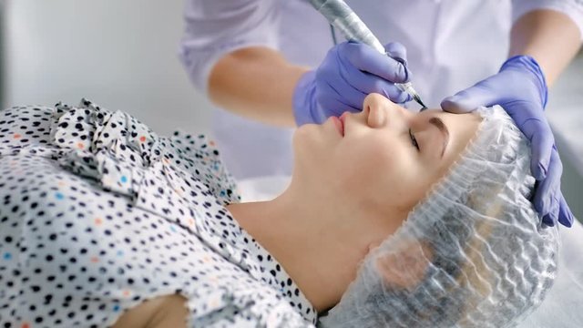 Beautician in gloves doing permanent makeup for eyebrows in beauty salon using tattoo pen machine