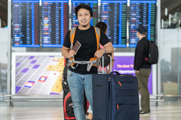 Portrait of Asian traveler with luggage with passport standing over the flight board for check-in at the flight information screen in modern an airport, travel and transportation concept.