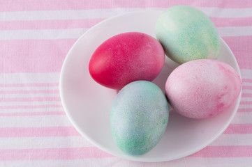 four multicolored painted Easter eggs on a white plate and  striped pink-white tablecloth. holiday easter greeting card, poster, wallpaper