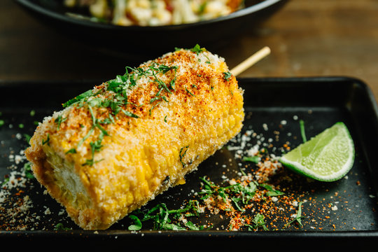 Mexican style grilled corn topping with cheese and chili powder served with sliced lime in black plate. 