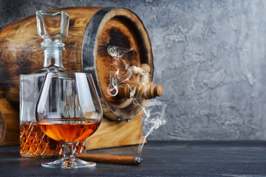 Strong alcoholic drink cognac in sniffer glass with smoking cigar, crystal decanter and vintage wooden barrel in cellar