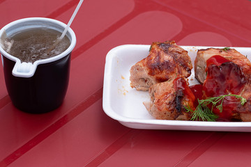 tea in a plastic glass and shish kebab in a rectangular plastic plate, a little green on a plastic table