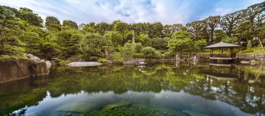 Central pond of Mejiro Garden which is surrounded by large flat stones under the foliage of the...